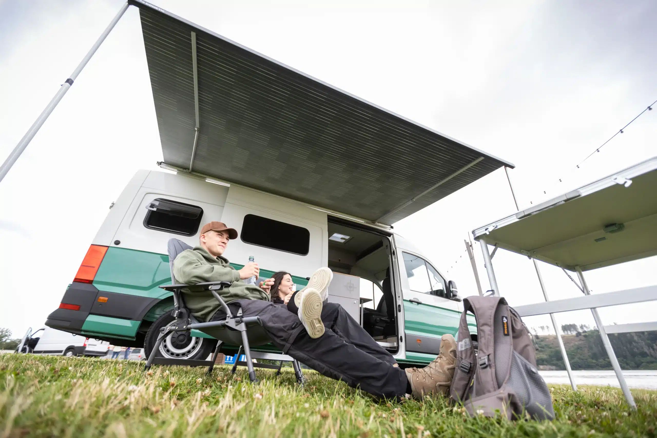Zwei Menschen entspannen vor einem geparkten WIGWAM Campervan unter einer Markise auf einer Wiese am See.