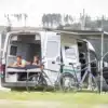 Offener WIGWAM Campervan (VW Crafter) mit Längsbett, Menschengruppe im Innenraum und Fahrrädern davor am Seeufer.