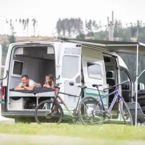 Offener WIGWAM Campervan (VW Crafter) mit Längsbett, Menschengruppe im Innenraum und Fahrrädern davor am Seeufer.