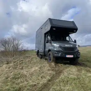 Das Allrad-Reisemobil Woelcke Autark Iveco XL 01 auf einer Wiese mit unebenem Untergrund unter wolkigem Himmel.