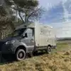 Woelcke Autark runner R10 Allrad-Wohnmobil, naturfarben, Mercedes Sprinter-Basis, auf einer Wiese vor Bäumen, Offroad-Reisemobil