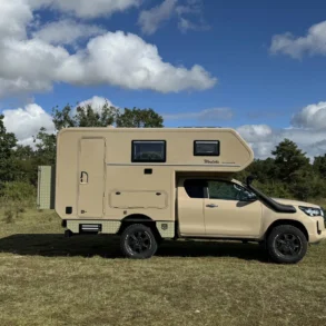 Seitenansicht des Woelcke Toyota Hilux Extra Cap 4x4 Camper auf einer Wiese unter bewölktem Himmel.
