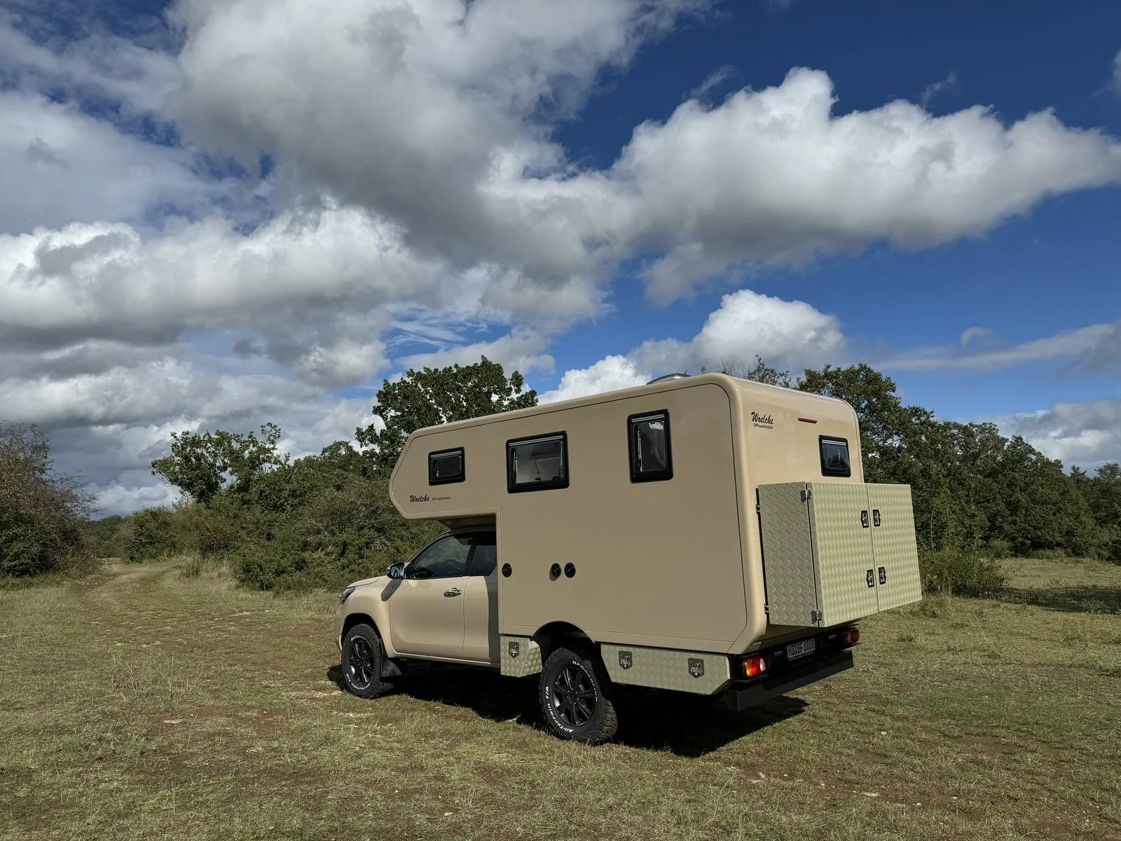Woelcke Toyota Hilux Extra Cap 4x4 Camper geparkt auf einer Wiese unter bewölktem Himmel.