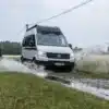 Carado CV595 X-EDITION Campervan auf VW-Basis fährt durch eine Wasserpfütze über eine nasse Wiese.
