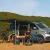 Fiamma F40 Van Dachmarkise in Deep Black und Royal Grey am Dach eines VW T6, ausgefahren, darunter zwei Camper mit Fahrrädern neben dem Bus.