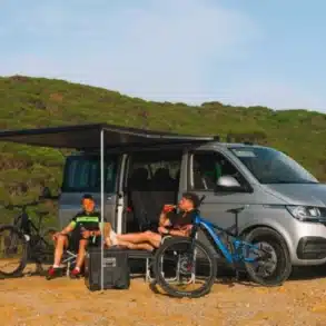 Fiamma F40 Van Dachmarkise in Deep Black und Royal Grey am Dach eines VW T6, ausgefahren, darunter zwei Camper mit Fahrrädern neben dem Bus.