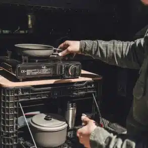Mann benutzt eine mobile Outdoor-Küchenkiste in einer schwarzen 48l Transformer Box mit Kocher und Kochgeschirr beim Camping am Fahrzeugheck