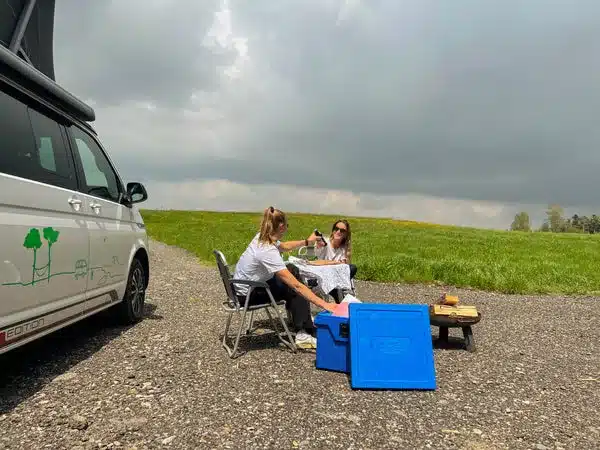 Zwei Frauen sitzen vor einem Campervan auf Stühlen an einem Tisch und nutzen eine blaue QOOL Box im Freien.