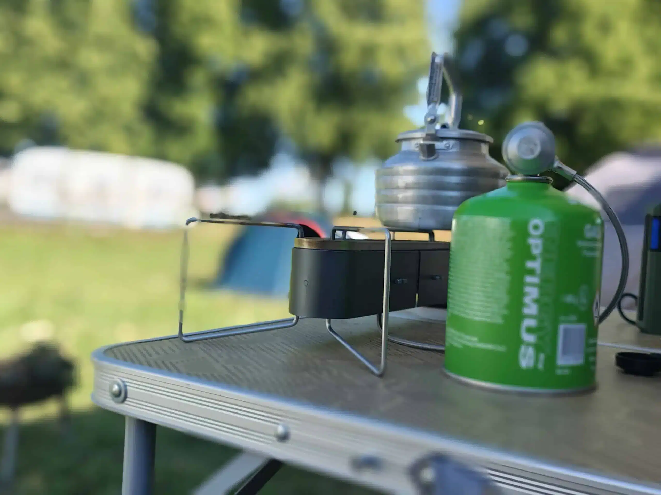 Optimus Gemini II Doppelbrenner Campingkocher mit angeschlossener Gaskartusche und Teekessel auf einem Campingtisch im Freien.
