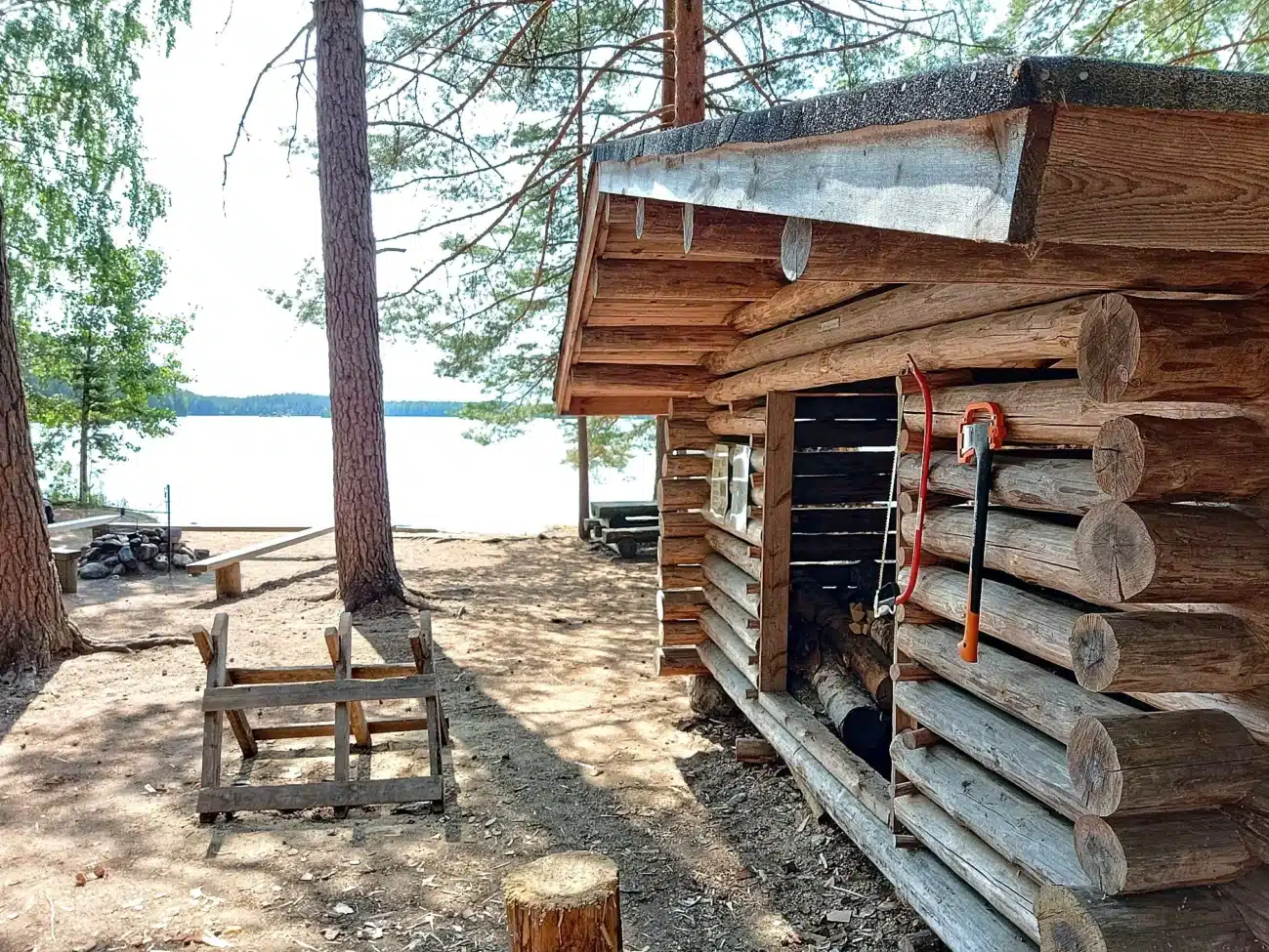 Blockhütte aus Holz an einem Seeufer mit Feuerstelle und Sitzgelegenheiten unter Bäumen.