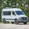 Außenansicht des Westfalia James Cook 600 E Camper-Vans auf Mercedes Sprinter Basis, stehend auf einem Parkplatz vor Bäumen.