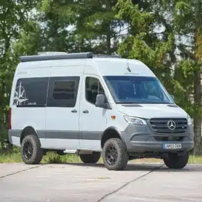 Außenansicht des Westfalia James Cook 600 E Camper-Vans auf Mercedes Sprinter Basis, stehend auf einem Parkplatz vor Bäumen.