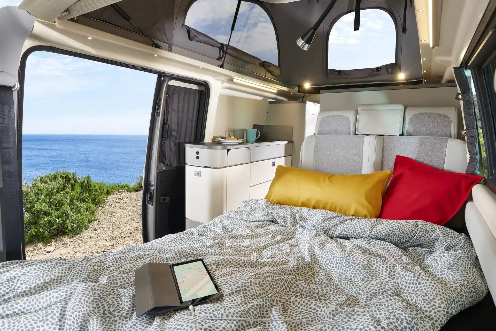 Der Innenraum des Westfalia Kepler One Campervans mit aufgeklapptem Bett, Kissen, Küchenschränken und geöffneter Schiebetür mit Blick auf das Meer.