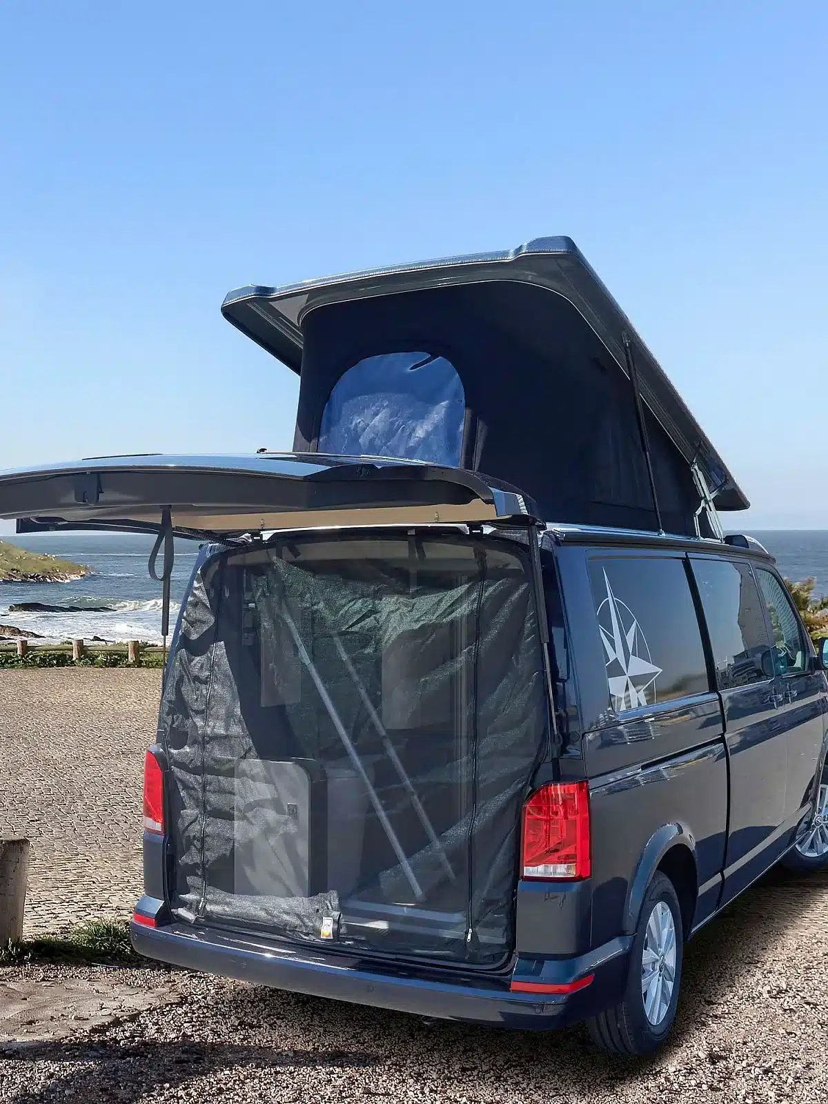 Der Westfalia Kepler One Campervan mit geöffnetem Aufstelldach und Heckklappe, geparkt an einem Küstenort.