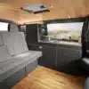 Innenansicht eines Campervans mit Holzdecke, grauer Sitzbank, Küche mit Edelstahlgaskochfeld und Stauraumlösungen.