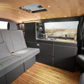 Innenansicht eines Campervans mit Holzdecke, grauer Sitzbank, Küche mit Edelstahlgaskochfeld und Stauraumlösungen.