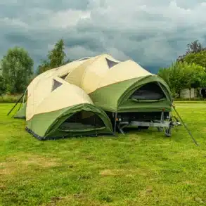 Geöffneter Alpenkreuzer OpenAir Faltcaravan auf einer grünen Campingwiese unter wolkigem Himmel.