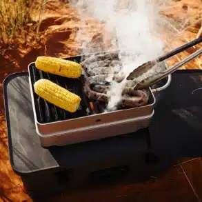 PACE® Camping-Kocher und Grill, belegt mit Würstchen und Mais, im Einsatz auf sandigem Untergrund.