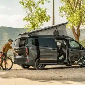 Panama Peak 5,45 Meter Campervan mit geöffneten Türen und Aufstelldach, zwei Fahrräder und eine Person davor, Natur im Hintergrund.