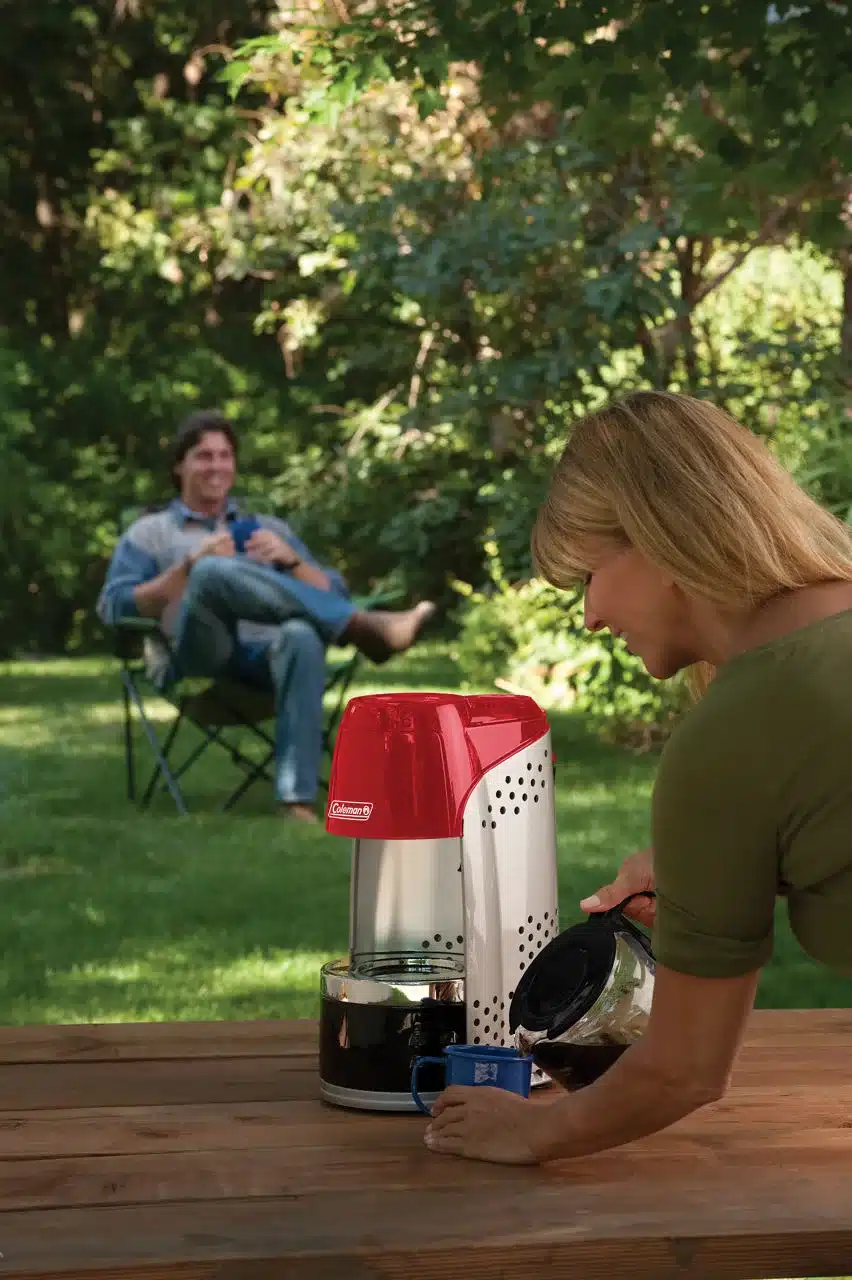 Frau gießt Kaffee aus der Glaskanne des Coleman QuikPot™ Propane Coffeemaker in eine Tasse beim Campen im Freien