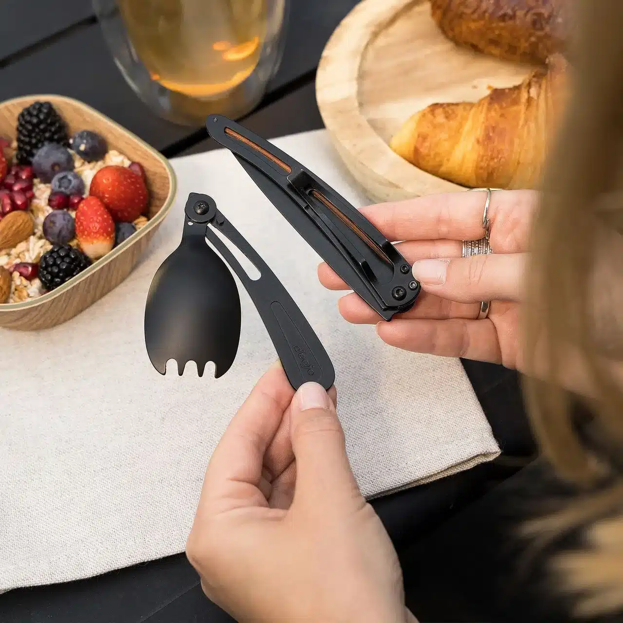 Schwarzer Spork Deejo 39g und Deejo 37g Messer werden von einer Person gehalten, Frühstück mit Obst, Croissant und Getränk auf einem Tisch im Hintergrund.