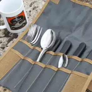 Campingbesteck organisiert in einer aufgerollten Tool Roll Hungry Edition aus Nylon, daneben eine weiße Tasse, alles auf Felsuntergrund.