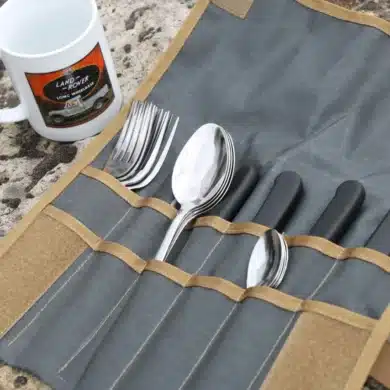 Campingbesteck organisiert in einer aufgerollten Tool Roll Hungry Edition aus Nylon, daneben eine weiße Tasse, alles auf Felsuntergrund.