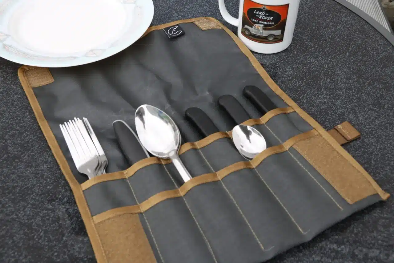 Campingbesteck in einer Rolltasche auf einem Tisch mit Teller und Becher, organisiert für mobile Küche.