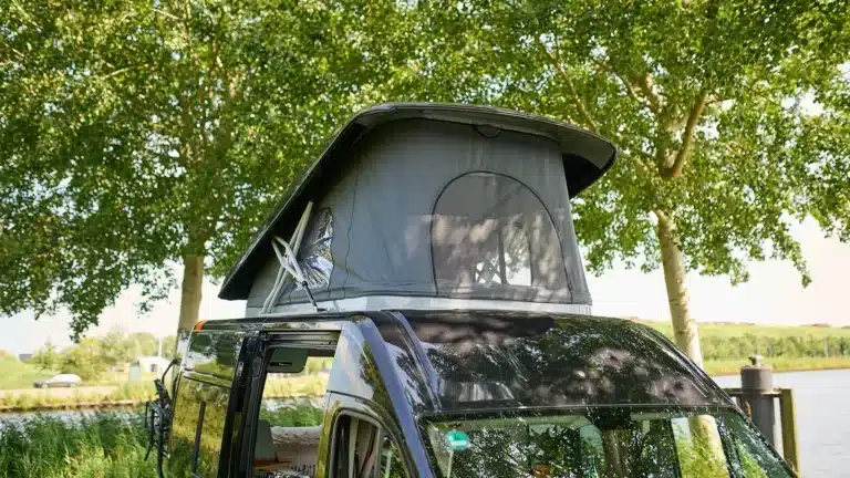 VAN-Jorn Family Camper mit geöffnetem Schlafdach unter Bäumen an einem Gewässer