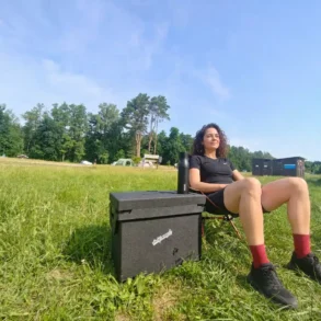 Frau sitzt entspannt neben einer Qool Box beim Camping auf einer Wiese.
