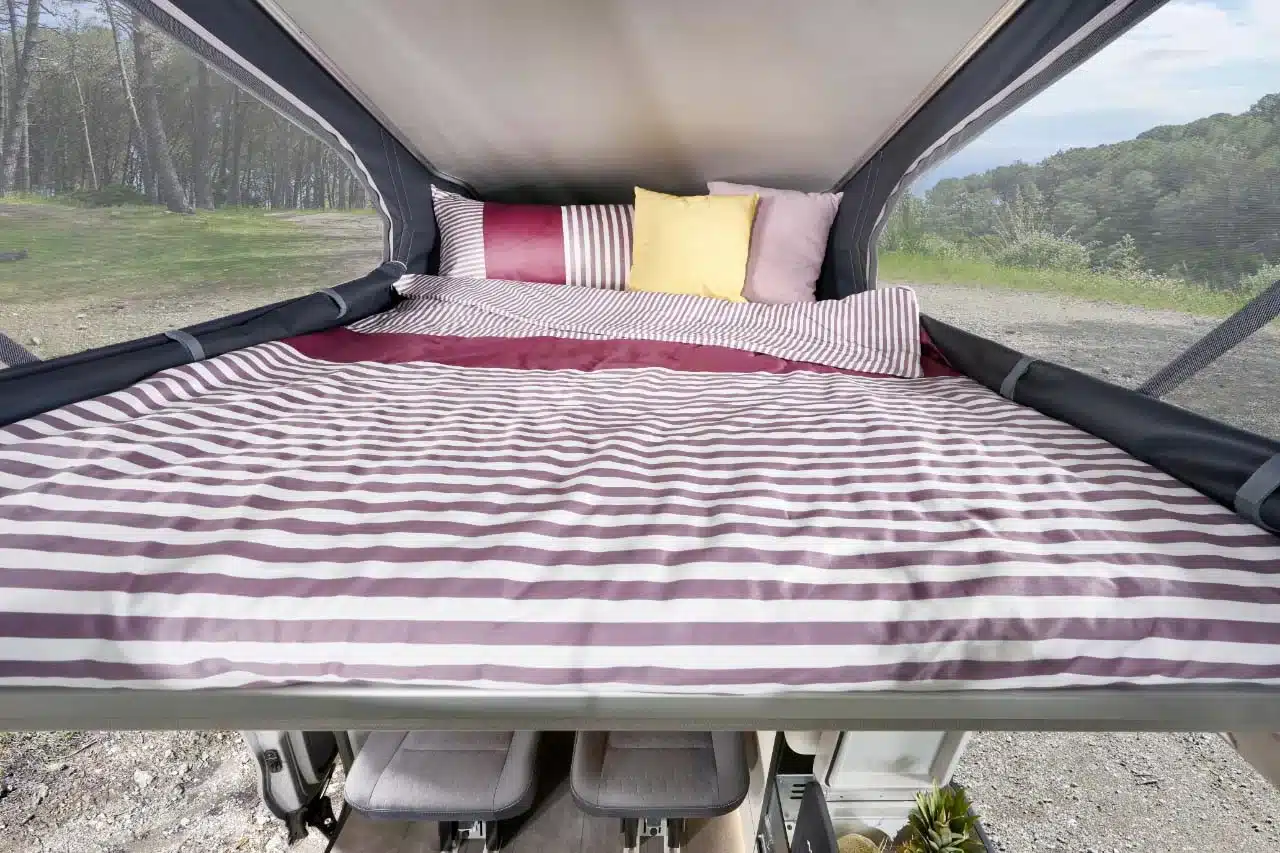 Blick in das Bett im Aufstelldach eines Westfalia Club Joker Urban mit gestreifter Bettwäsche und Kissen.