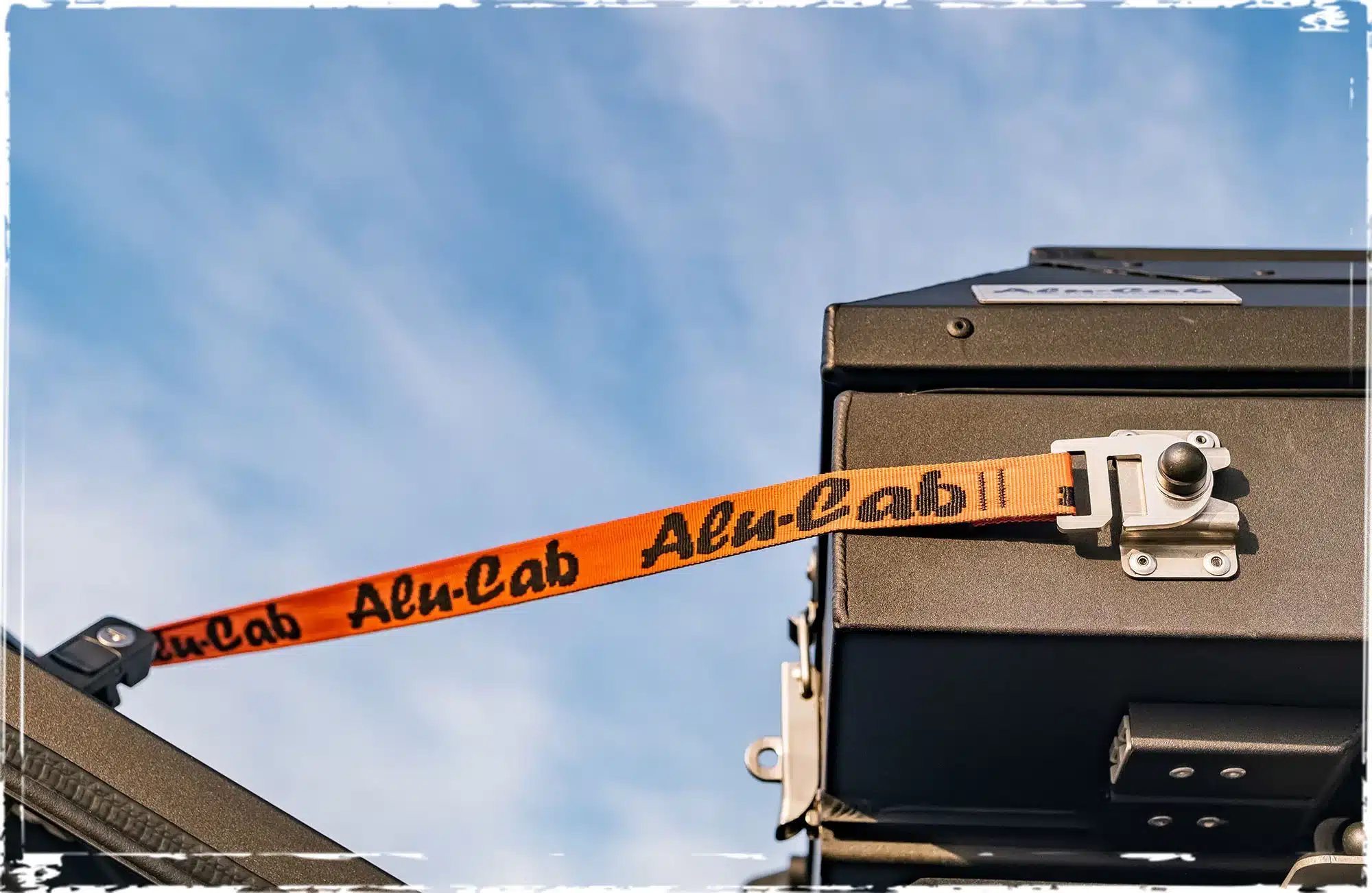 Detailansicht der Befestigung der Alu-Cab Markise 180°, rechts mit orangefarbenem Gurt am Fahrzeug bei blauem Himmel.