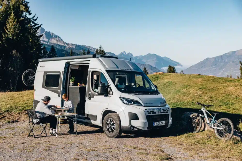 Weißer Sunlight CLIFF Adventure Camper Van mit geöffnetem Seiteneinstieg, Mountainbikes und Sitzgruppe vor einer alpinen Berglandschaft
