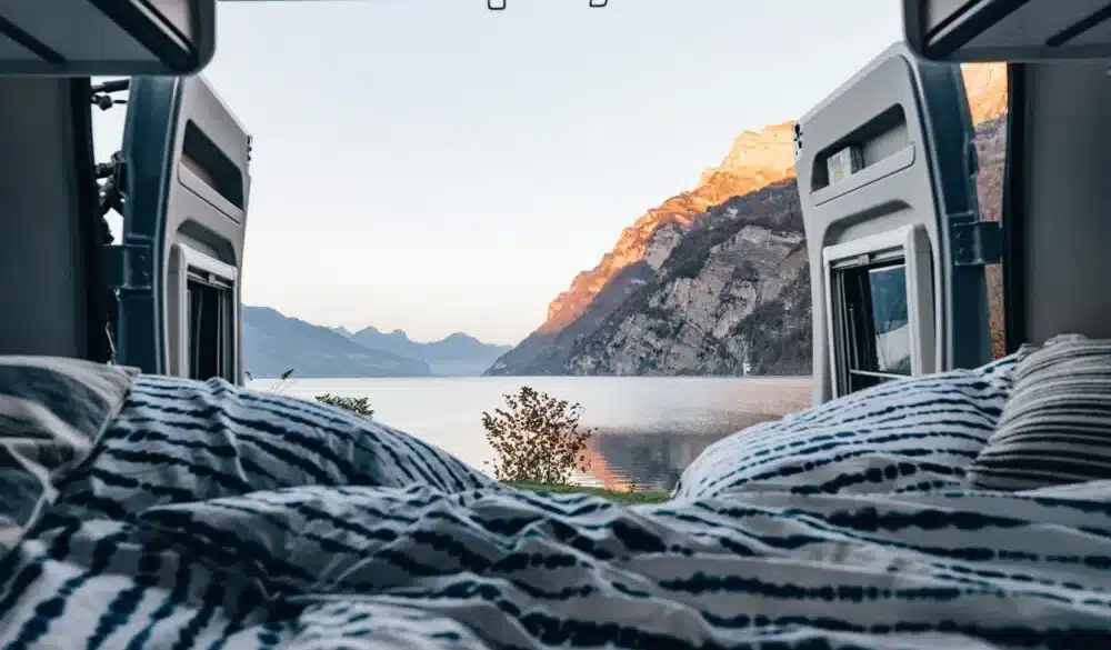 Bett im Innenraum eines Camper Vans mit geöffneten Hecktüren und Blick auf einen See und Berge