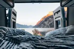 Bett im Innenraum eines Camper Vans mit geöffneten Hecktüren und Blick auf einen See und Berge