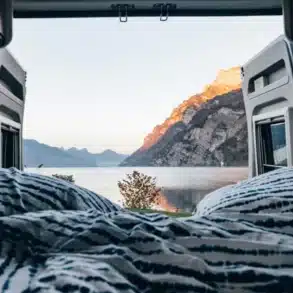 Bett im Innenraum eines Camper Vans mit geöffneten Hecktüren und Blick auf einen See und Berge