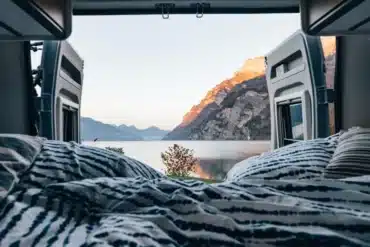 Bett im Innenraum eines Camper Vans mit geöffneten Hecktüren und Blick auf einen See und Berge