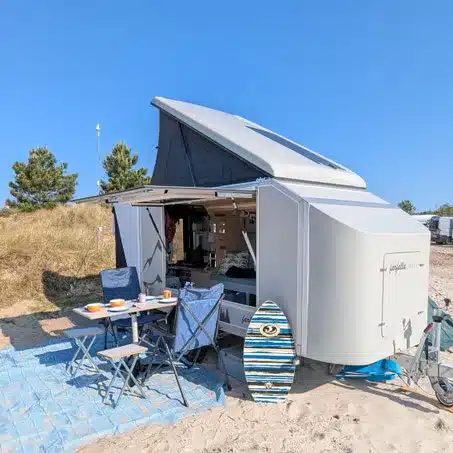 Farfalla Camper flip mit ausgeklappter Panoramaklappe, Campingmöbeln und Surfbrett am Strand.
