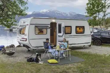 Weißer Fendt-Caravan APERO 515 mit geöffneter Tür an einem See vor einer Bergkulisse