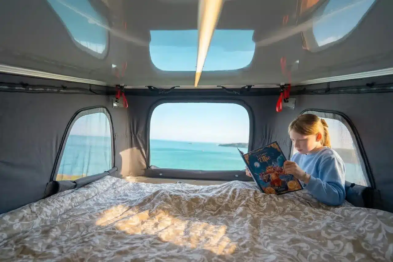 Kind im Flex 5 Confort Campervan liest im Hochbett des Aufstelldachs mit Ausblick auf das Meer