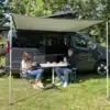 Campervan Flex Confort PLUS mit aufgestellter Markise, ausgeklappten Stühlen und Tisch, zwei Personen beim Essen, Fahrradträger am Heck.