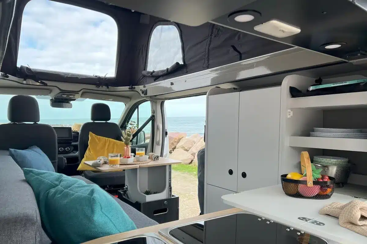 Campervan Flex Confort PLUS mit geöffnetem Aufstelldach, Sitzbereich, offener Küche und Blick aufs Meer.