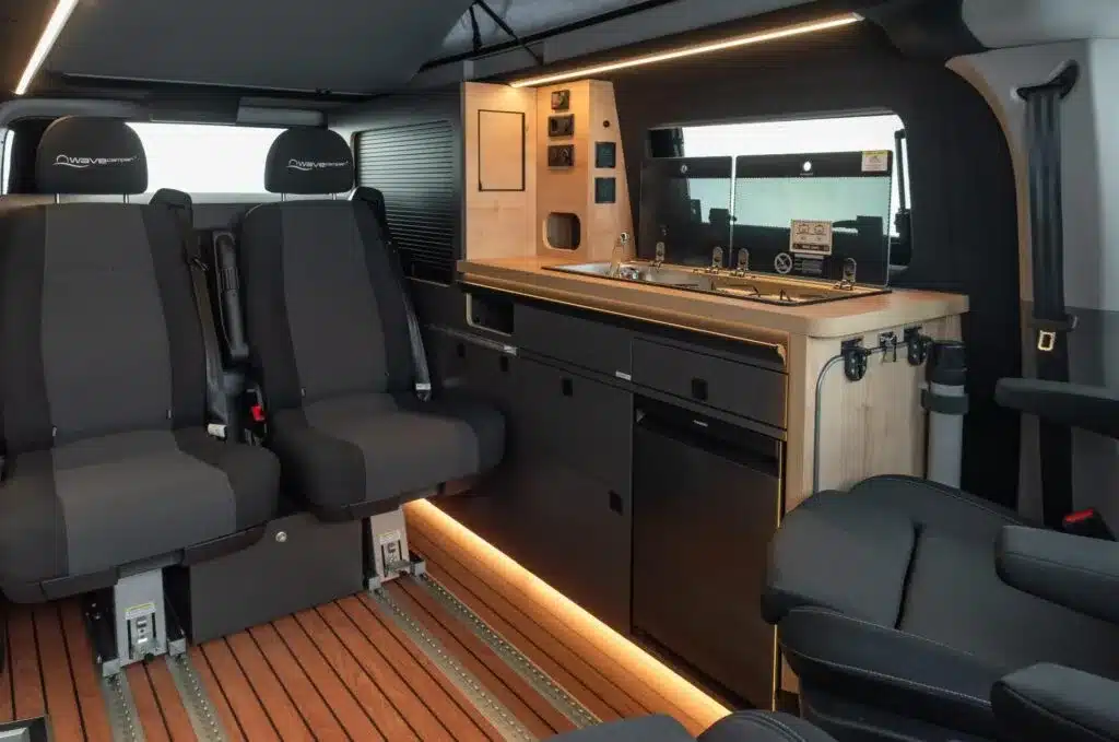 Innenansicht des Ford Custom 4x4 Wavecamper mit zwei einzelnen Sitzen auf Schienensystem und Küchenzeile mit Spüle, Herd und Kühlschrank
