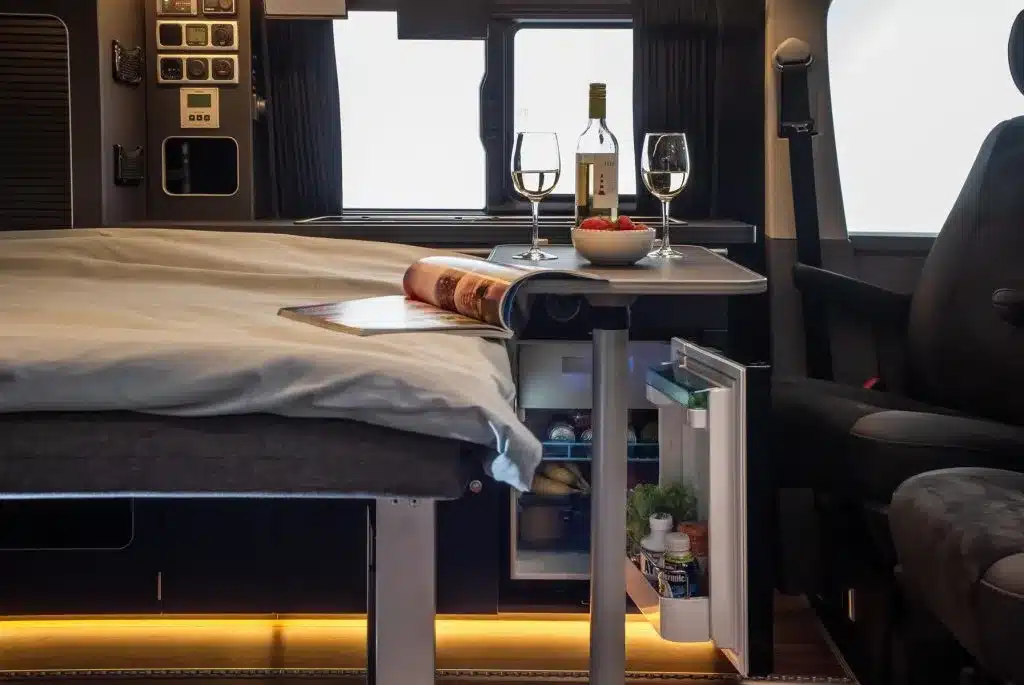 Blick in den Innenraum des Ford Custom 4x4 Wavecamper mit gedecktem Tisch, offenem Kühlschrank und Bett.