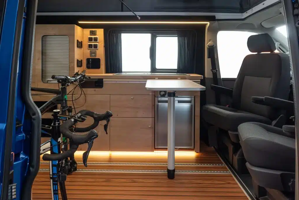 Innenraum des Ford Custom 4x4 Wavecamper mit Küchenblock, Tisch, Schienensystem und abgestelltem Fahrrad