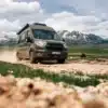 Olivgrüner Sunlight IBEX Ford Camper Van fährt auf einem steinigen Bergweg vor verschneiten Bergen.