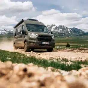 Olivgrüner Sunlight IBEX Ford Camper Van fährt auf einem steinigen Bergweg vor verschneiten Bergen.