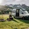 Sunlight IBEX VW 4x4 Camper steht auf einer Wiese vor Bergen, Campingzubehör und Lagerfeuer im Vordergrund