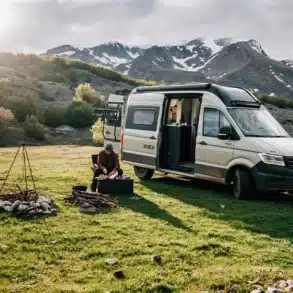 Sunlight IBEX VW 4x4 Camper steht auf einer Wiese vor Bergen, Campingzubehör und Lagerfeuer im Vordergrund