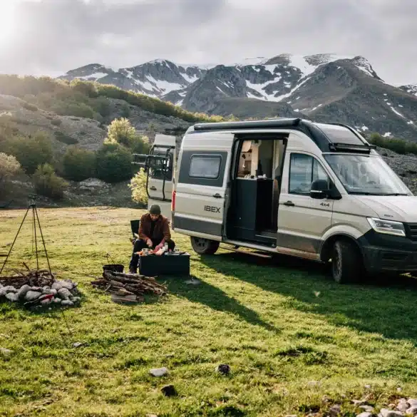 Sunlight IBEX VW 4x4 Camper steht auf einer Wiese vor Bergen, Campingzubehör und Lagerfeuer im Vordergrund